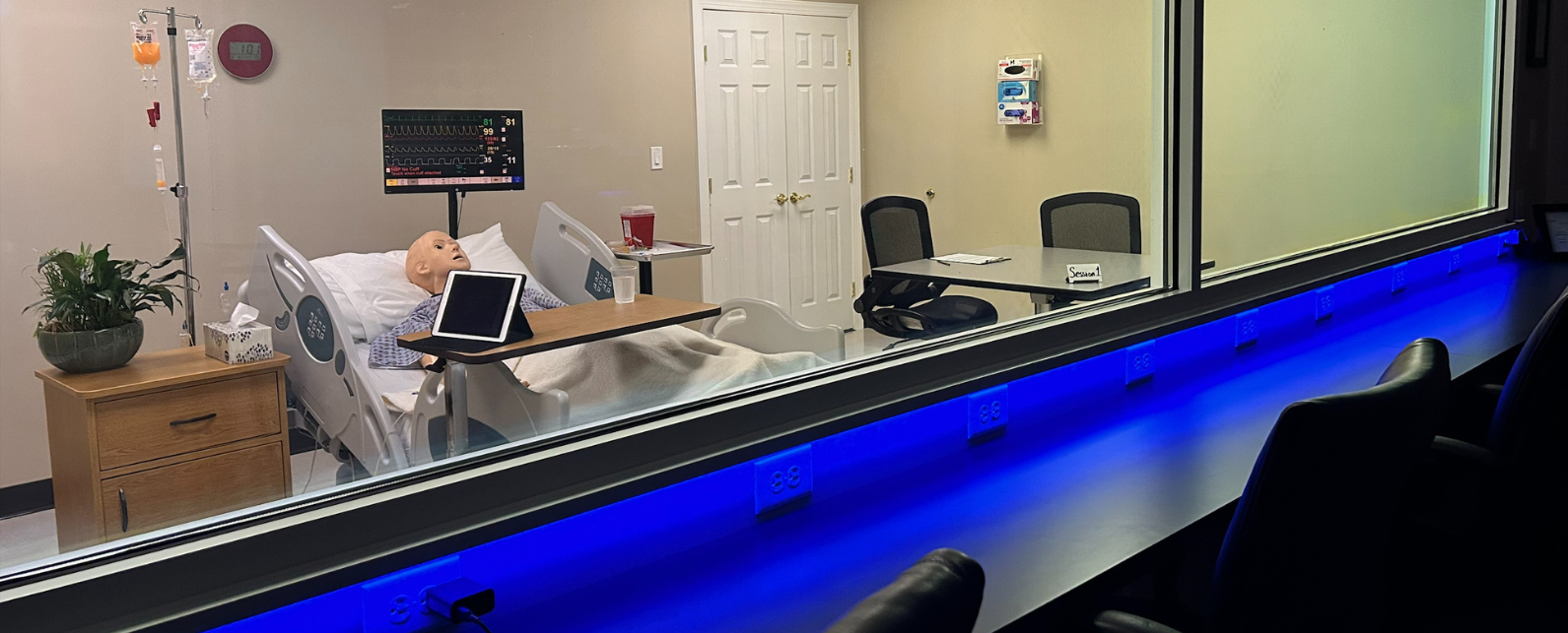 Chairs near a window to see a laboratory simulation of a mannequin in a hospital bed with a bedside table and infusion pole.