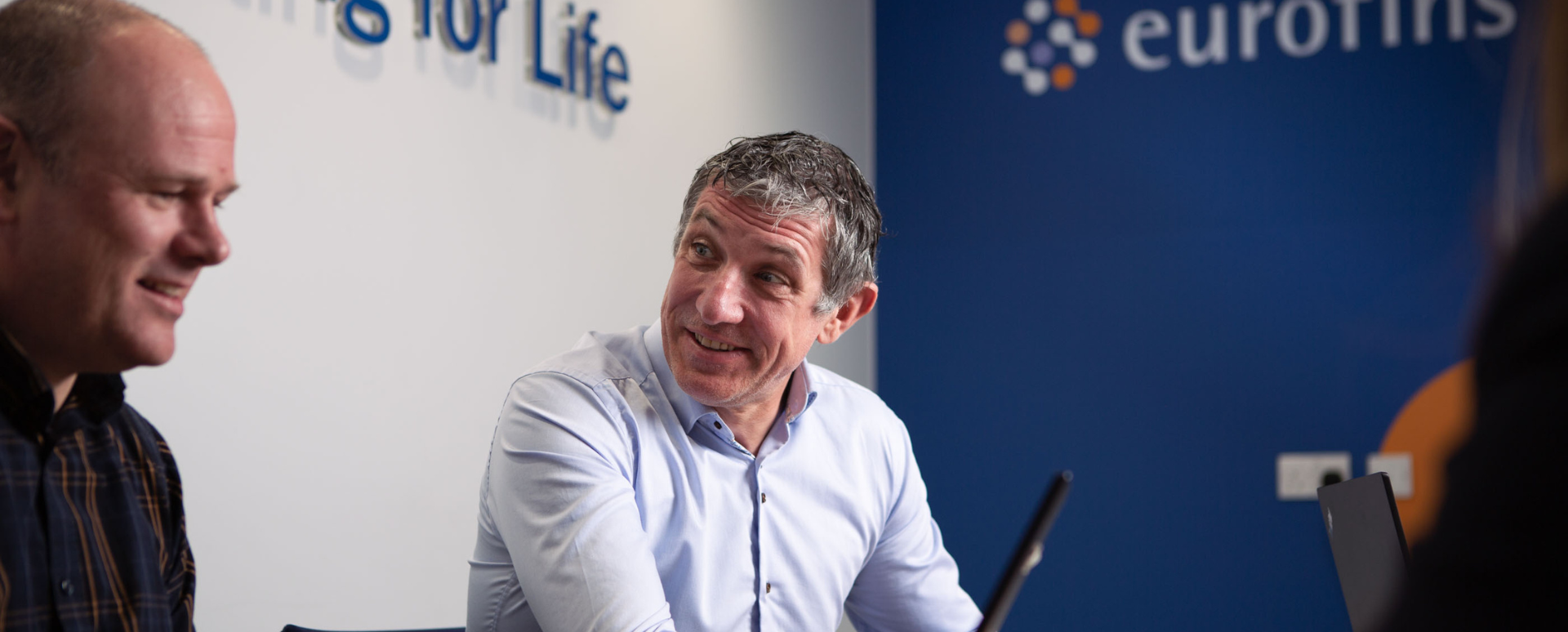 Deux consultants discutent dans un bureau moderne, le logo d'Eurofins étant visible à l'arrière-plan. L'image illustre un environnement professionnel collaboratif axé sur les dispositifs médicaux.