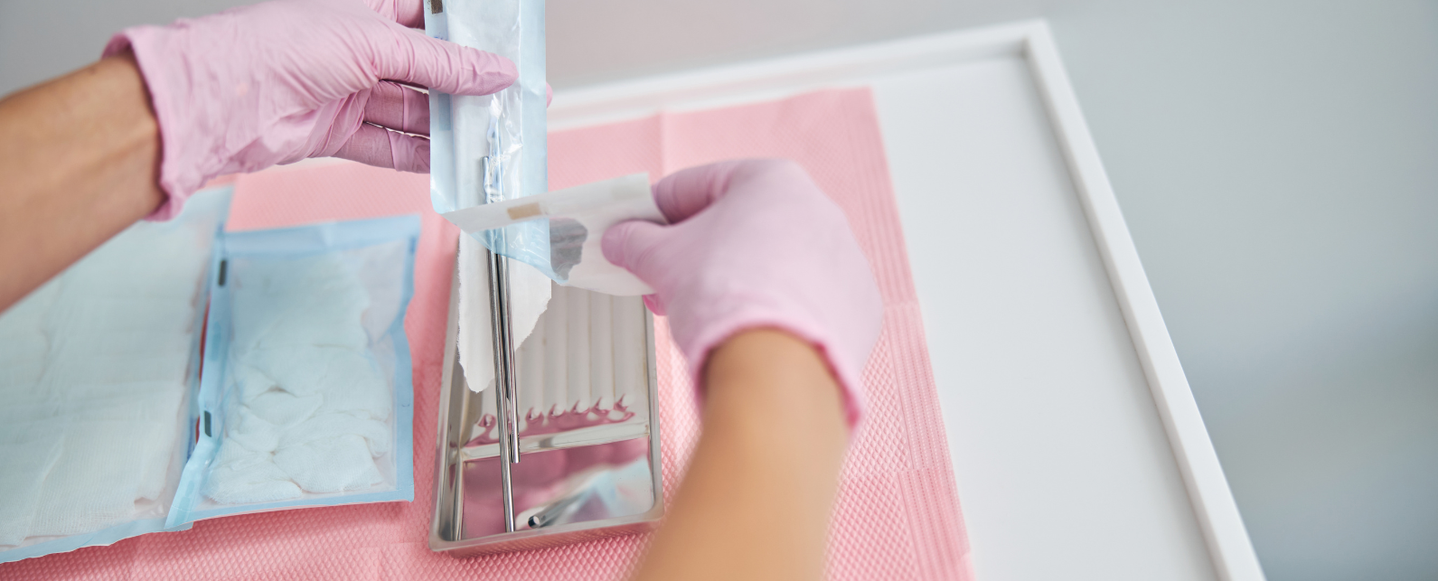 Deux mains gantées ouvrent un emballage en plastique contenant un scalpel sur une table blanche recouverte d'un drap de protection rose.