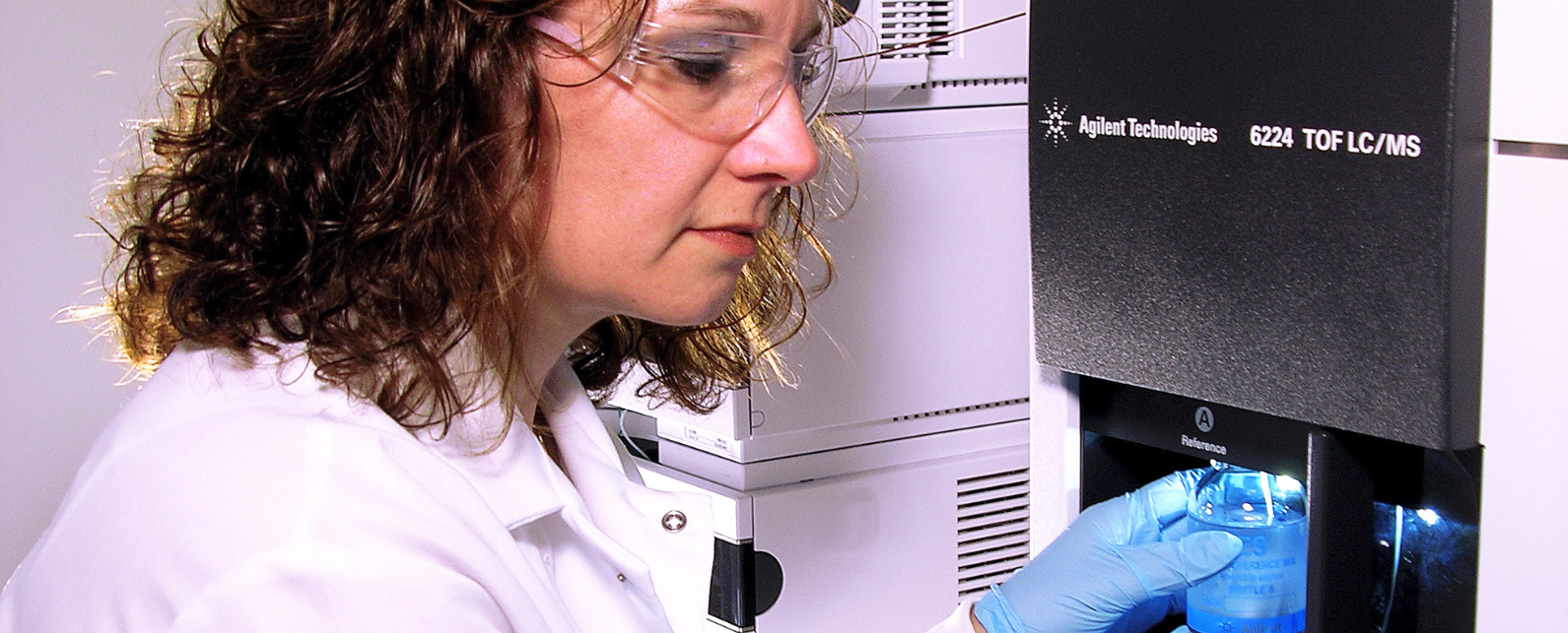Une femme brune en blouse et lunettes tient un échantillon sur un LC/MS Agilent 6224 TOF avec des mains gantées.